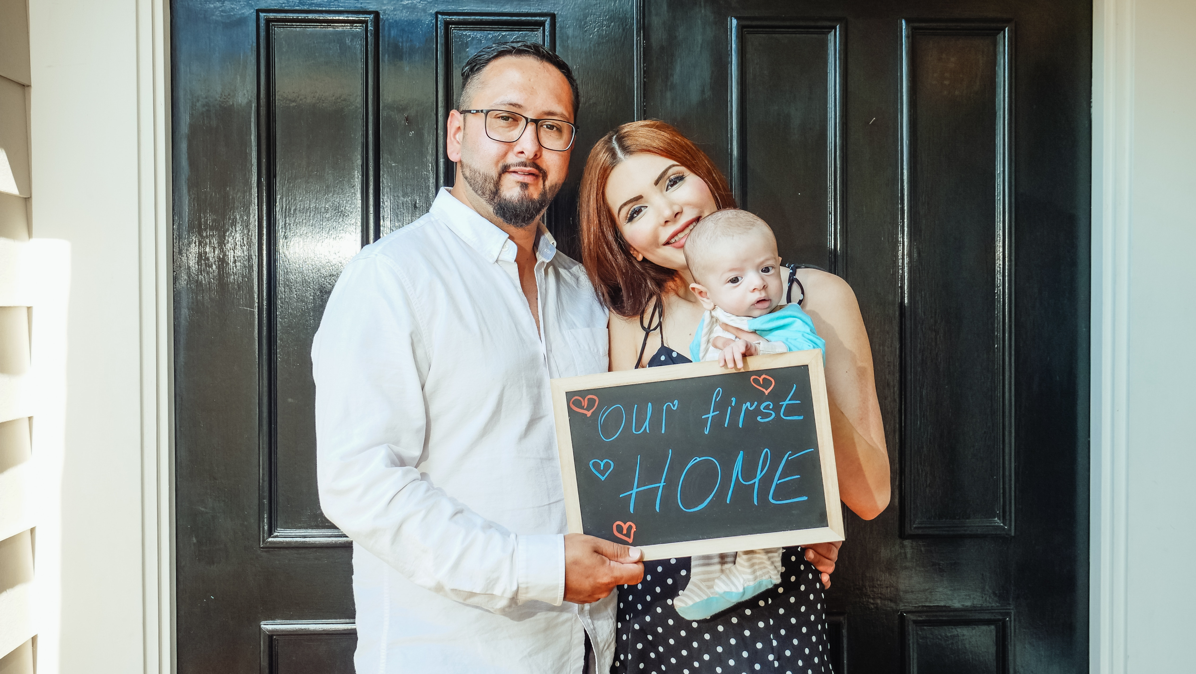 A man with glasses and a woman with red hair hold up an infant child, along with a sign that says "Our first home"
