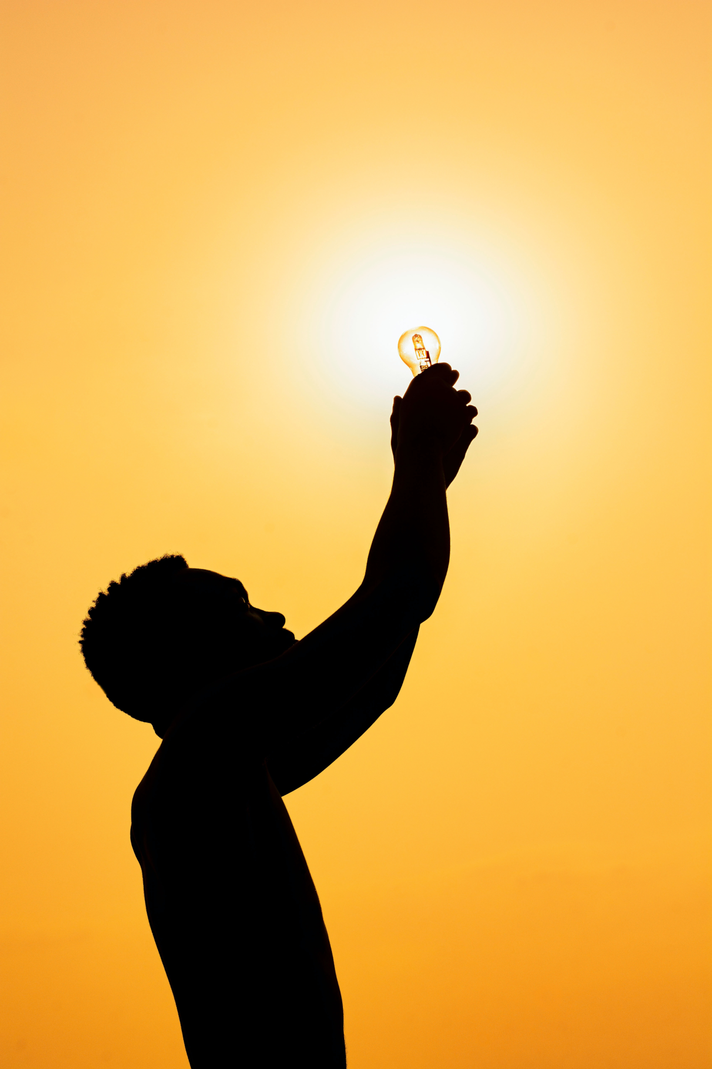 A man reaches towards a gold sky and cups a lightbulb in his hands, with the sun in the background.